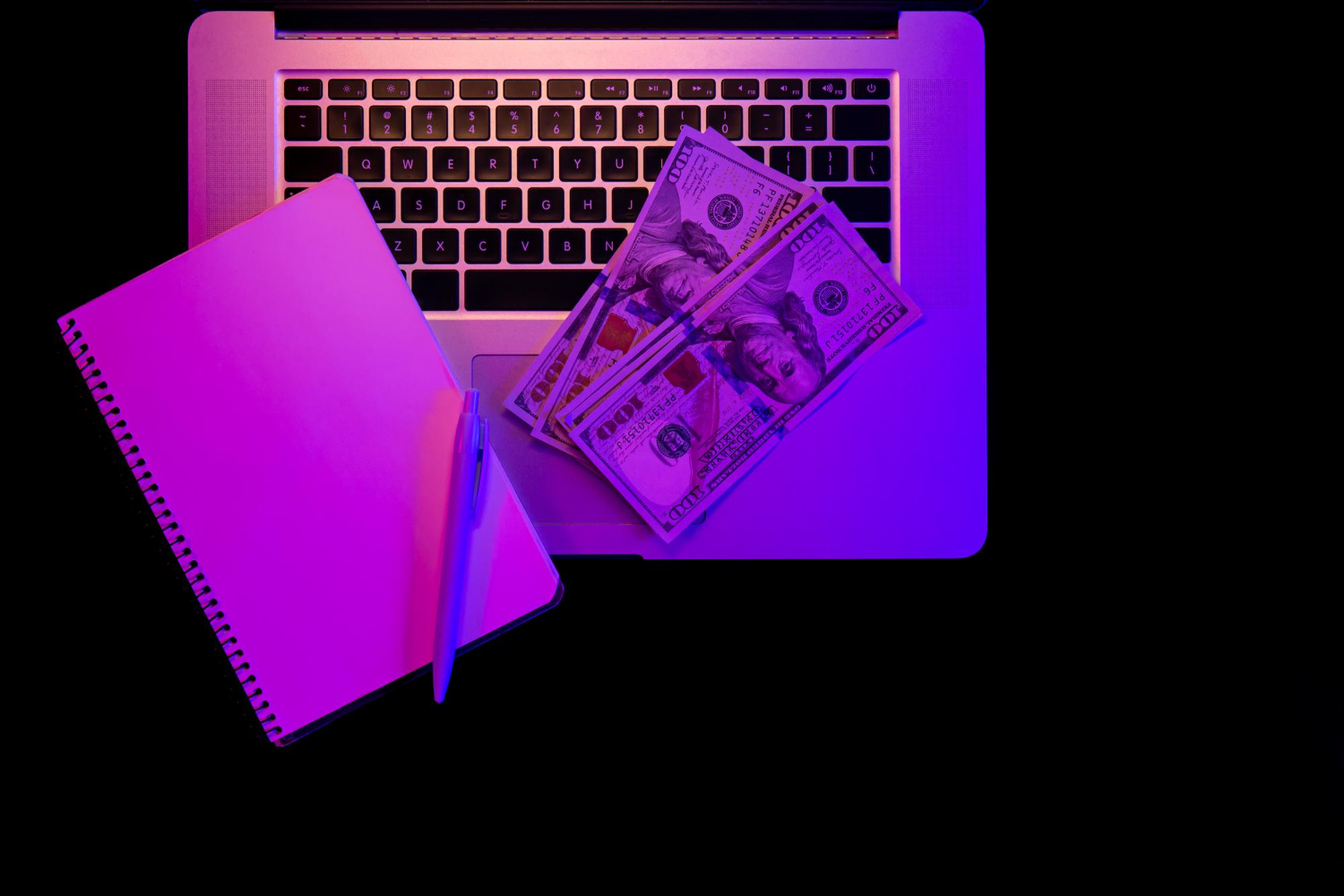 Laptop with a blank notepad and 100 dollar bills on top under neon light